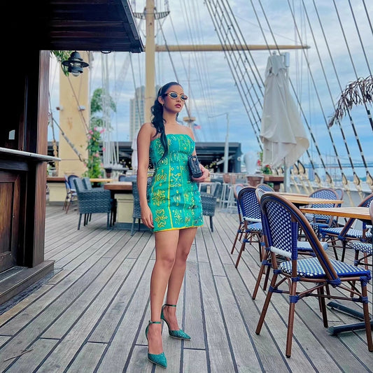 A woman stands on a ship's deck wearing a bright green strapless mini Habesha Wedding Dress with yellow floral patterns, holding a black handbag. The background features a large sailing ship mast, tables, and chairs on the deck.