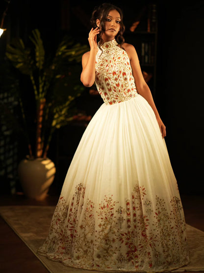 A woman models a sleeveless high-neck ball Habesha Wedding Dress  with a cream tulle skirt and a bodice decorated with red and gold floral embroidery, set against a dark, softly lit background with a potted plant.