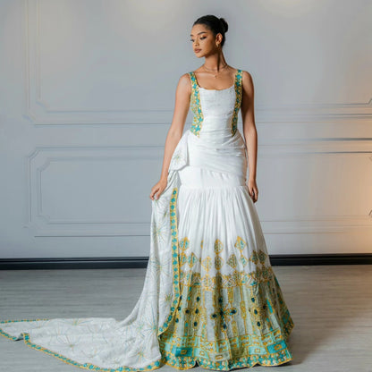 A woman models a white sleeveless traditional Habesha Wedding Dress with teal and gold embroidered borders, standing against a light gray paneled wall.