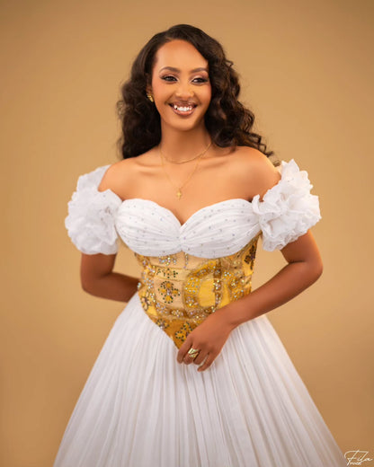 A smiling woman wearing an off-the-shoulder white Habesha Wedding Dress  with a gold beaded corset bodice and a full white skirt, posed against a warm beige studio background.