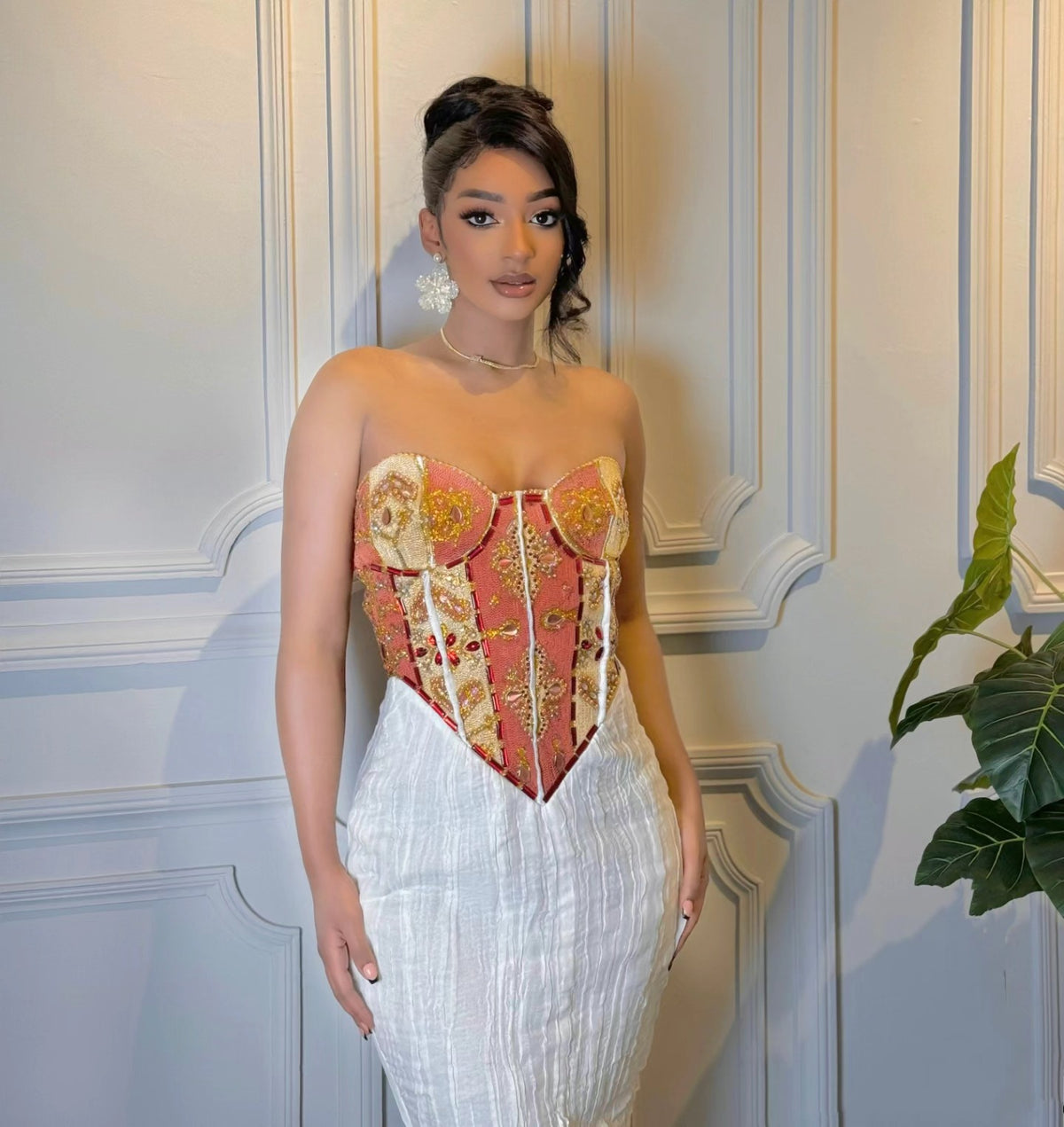 A woman models a strapless corset-style Habesha wedding dress with a red, gold, and beige brocade pattern, paired with a white textured skirt, standing against a white paneled wall.