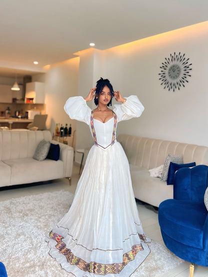 A woman modeling a white Ethiopian Dress with voluminous puff sleeves, a fitted bodice, and colorful geometric patterned borders, standing in a modern living room.
