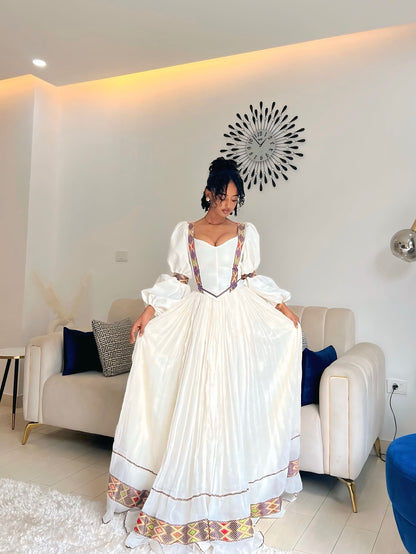 A woman wearing a long Ethiopian Dress with colorful geometric patterned trim, standing in a modern living room with a beige sofa and warm wall lighting.