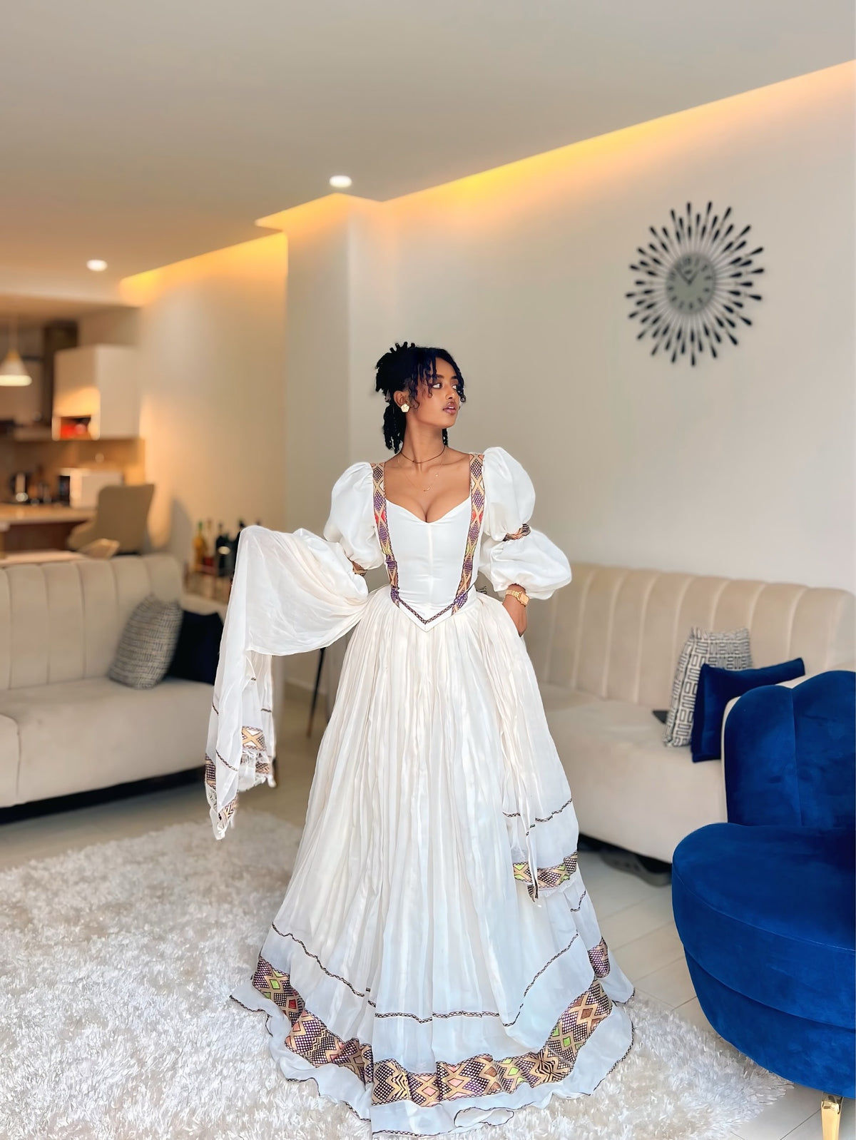 A woman wearing a white Ethiopian Dress with ornate gold, red, and blue embroidery, standing in a modern living room with beige sofas and a blue accent chair.