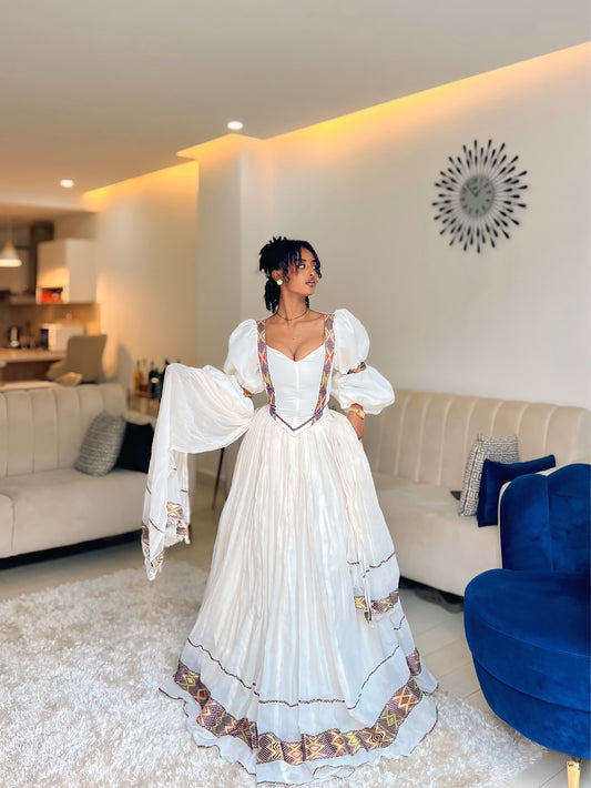 A woman wearing a white Ethiopian Dress with ornate gold, red, and blue embroidery, standing in a modern living room with beige sofas and a blue accent chair.