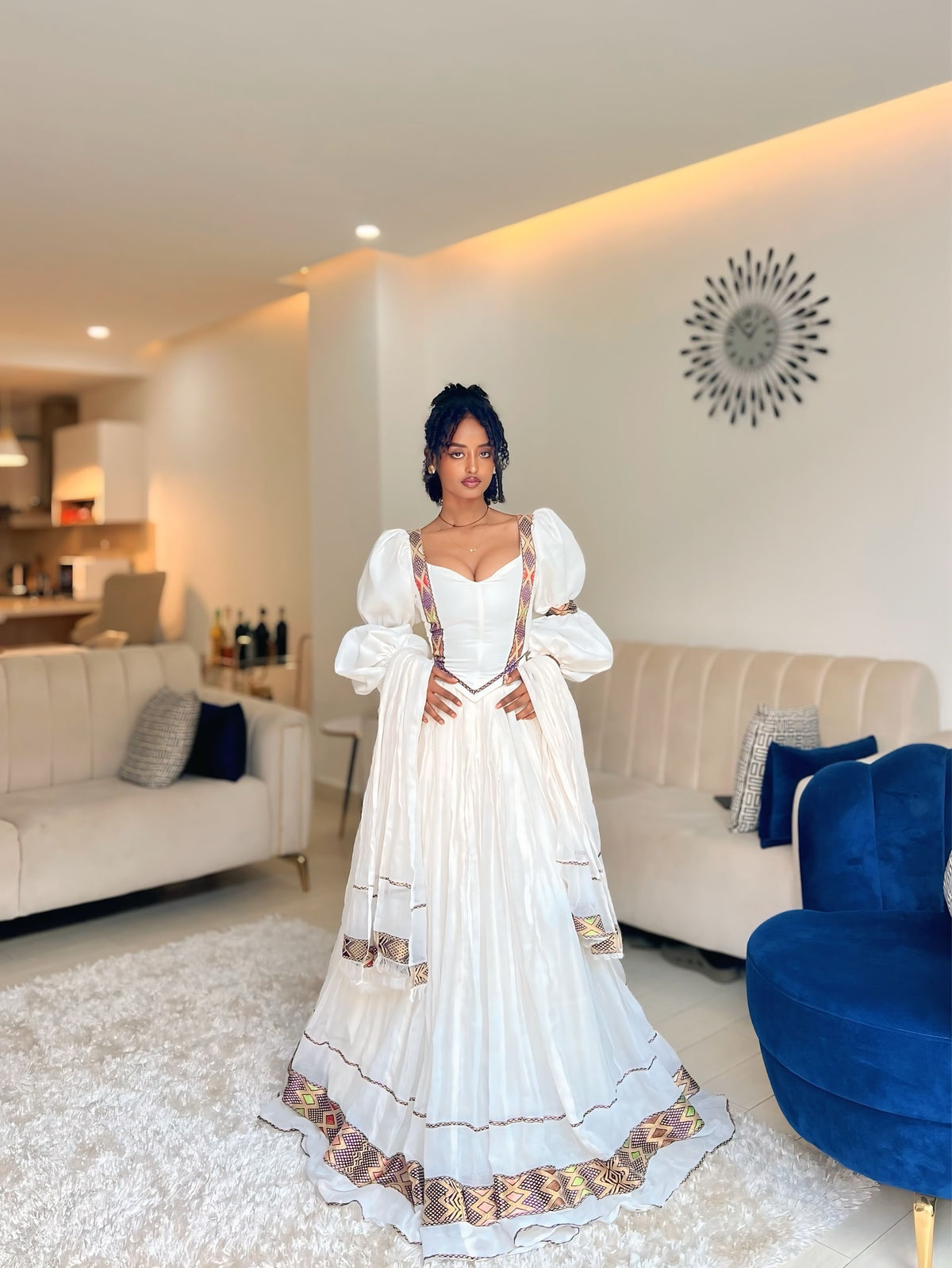 A woman wearing a long white Ethiopian Dress with gold and red patterned borders, standing in a modern living room with beige sofas and a blue accent chair.