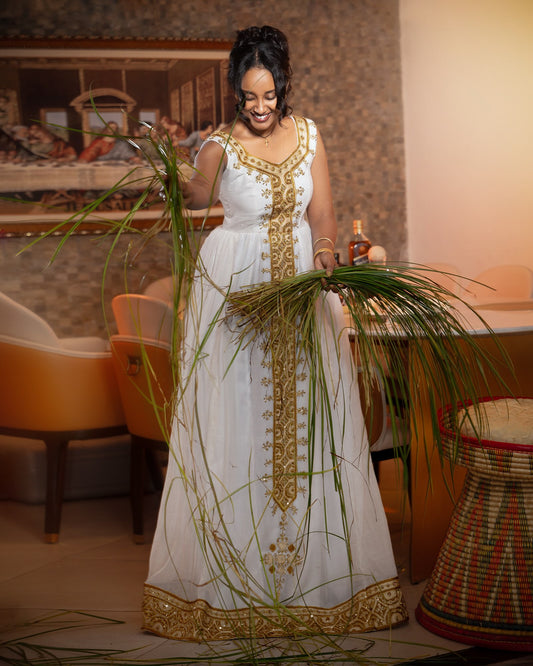 A woman wearing a white sleeveless Ethiopian Store with ornate gold embroidery and a gold border stands indoors while holding tall green grasses. The room has warm lighting, upholstered chairs, and a woven basket on the right.