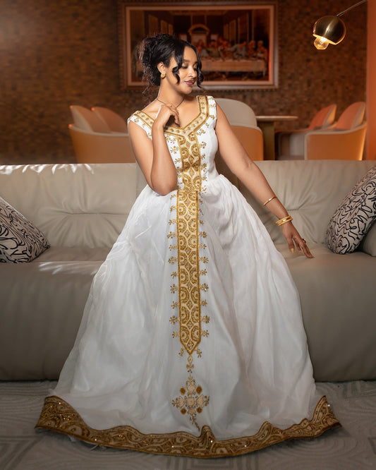 A woman wearing a white sleeveless Ethiopian Store with ornate gold embroidery and a flared skirt, posed in front of a beige sofa in a warmly lit indoor setting.