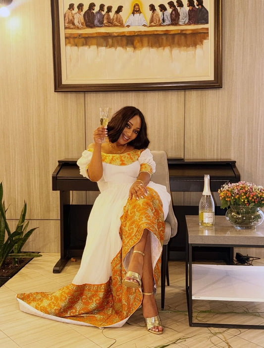 A woman seated in a chair indoors, wearing a white Ethiopia clothes Traditional with a bright orange and yellow patterned yoke and hem, holding a glass. A framed painting hangs on the wall behind her, with a piano, side table, flowers, and a bottle on the table visible in the room.