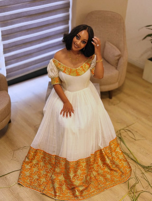 A woman wearing a white off-the-shoulder maxi Ethiopia clothes Traditional with gold and orange embroidered details, posing in a modern indoor setting with wooden floors and beige armchairs.