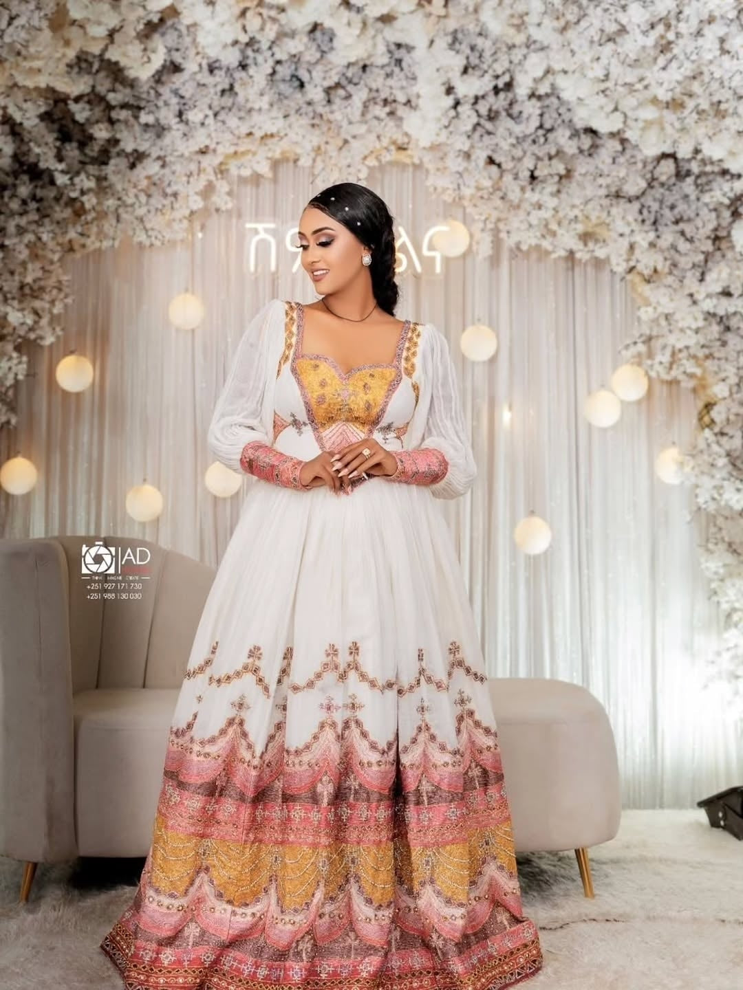 Woman in a white and colorful Ethiopian Habesha Couple Set standing in a decorated room with flowers and lights.