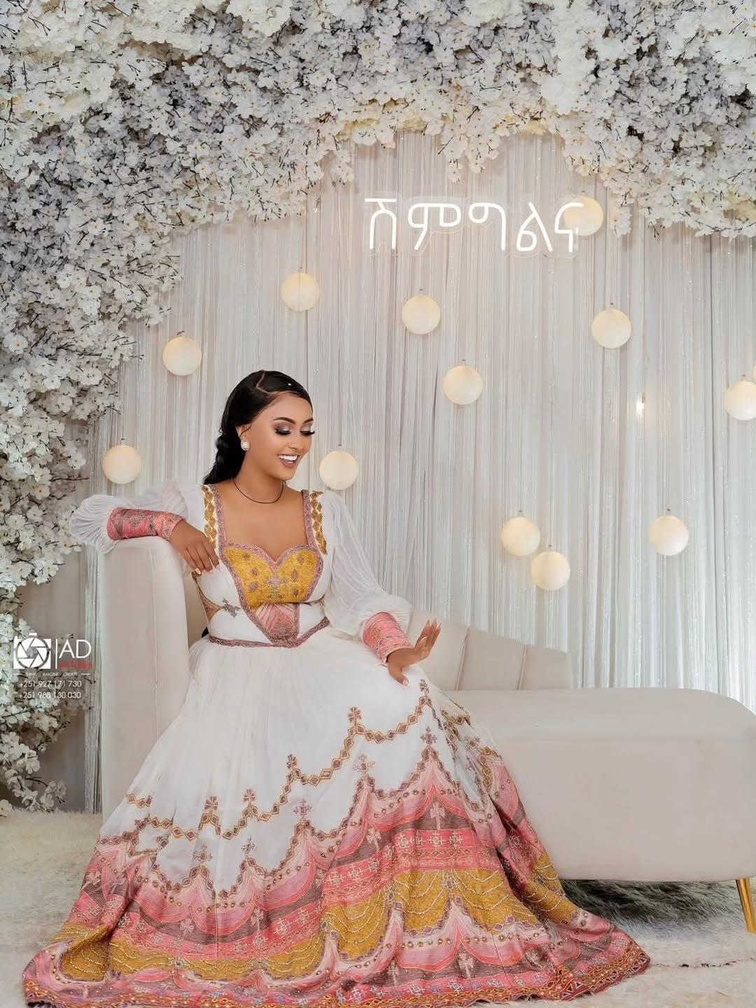 Woman in a colorful Ethiopian Habesha Couple Set sitting on a couch with floral decorations and lights in the background