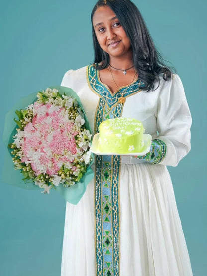 Woman in traditional Green Habesha Kemis holding a bouquet of flowers and a cake against a teal background
