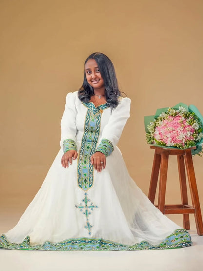 Woman in a white dress with green and blue patterns standing next to a bouquet of flowers on a wooden stool against a beige background.