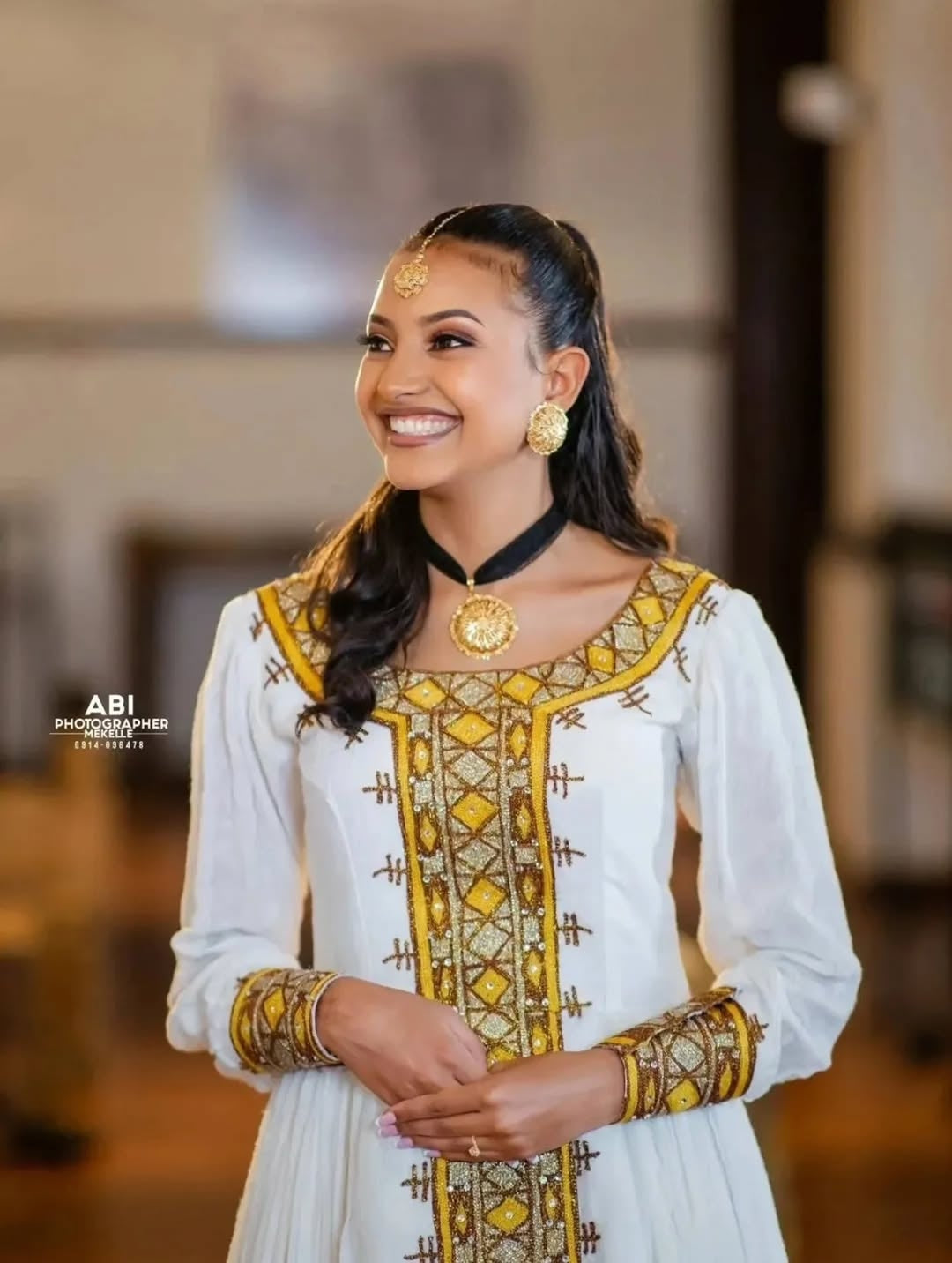 Woman in traditional attire with gold and yellow patterns, smiling indoors.