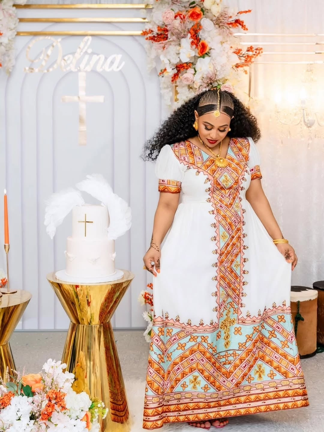 Woman in a colorful Habesha dress standing in front of a decorated arch with floral arrangements.
