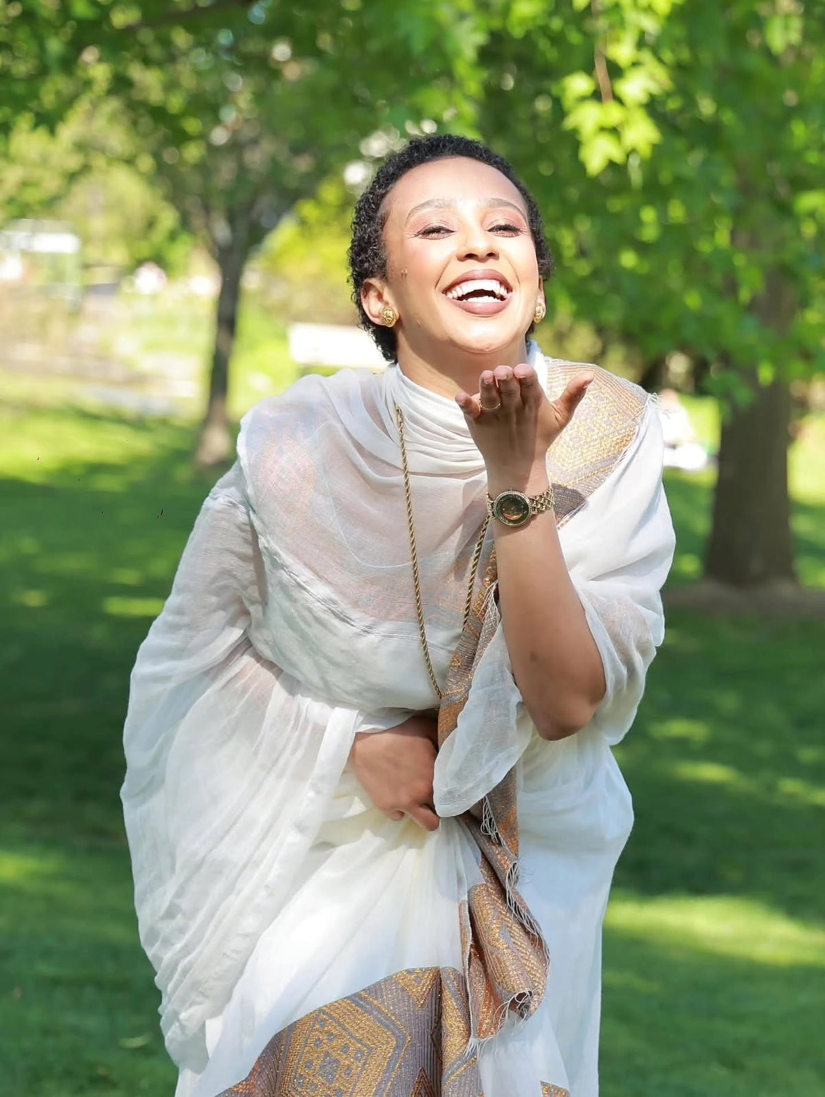 Woman in a white Habesha Kemis  with a gold border standing outdoors in a park.