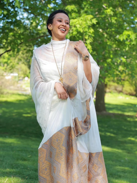 Woman in a white and gold traditional Habesha Kemis  standing outdoors with greenery in the background
