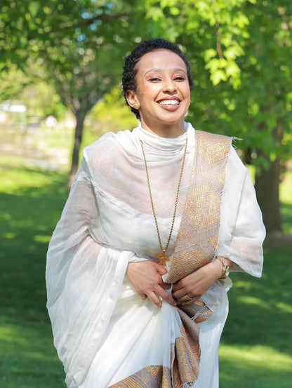 Woman in a white and gold traditional outfit standing outdoors with greenery in the background