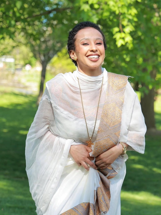 Woman in a white and gold traditional outfit standing outdoors with greenery in the background
