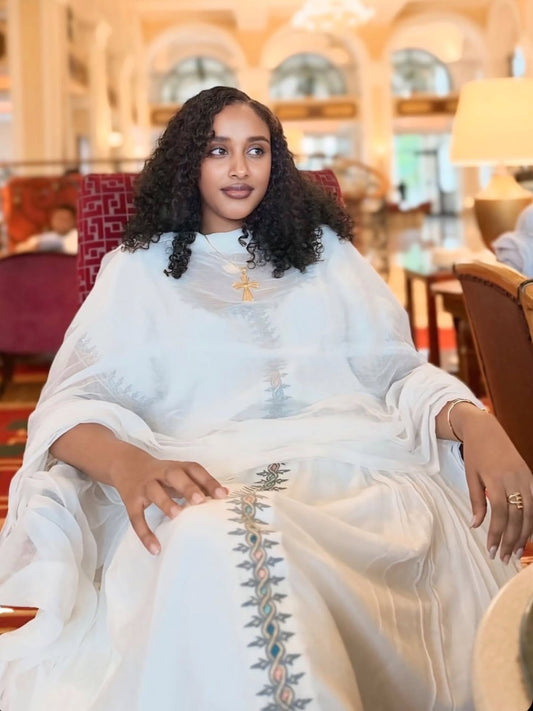 A woman with curly black hair sits in a grand indoor setting wearing a white traditional habesha kemis with delicate embroidery and a gold cross necklace.