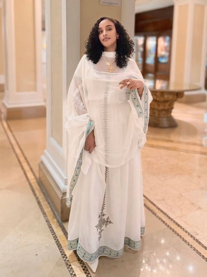 A woman wearing a white traditional habesha kemis with a matching sheer outer layer and light green patterned borders, standing in a grand indoor hallway with beige columns and tiled floors.