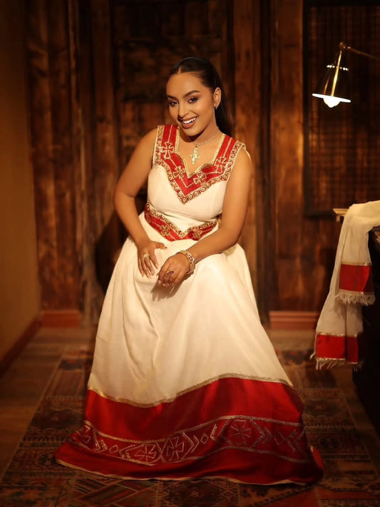 A woman wearing a traditional white Habesha Zuria with red embroidery and a red hem, standing in a warmly lit indoor setting.
