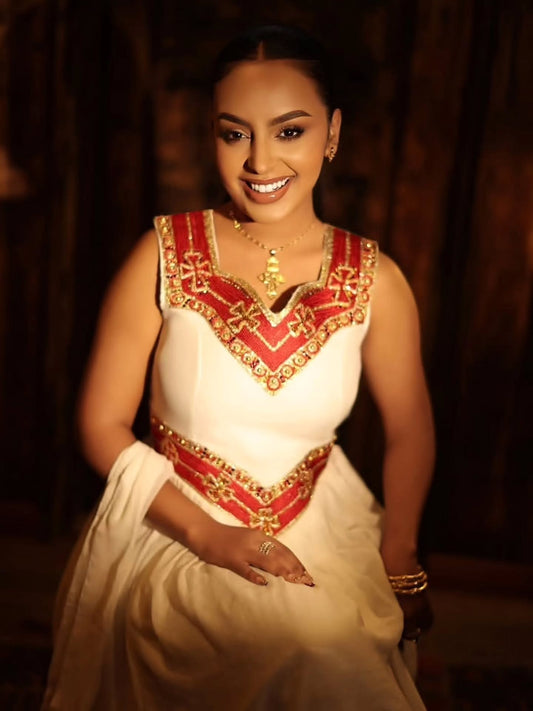 A woman wearing a traditional white Habesha Zuria with red and gold embroidered details, seated against a dark, warm-toned background.