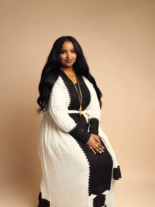 A woman modeling a long white textured robe with black Habesha Kemis waist tie, posed against a solid beige studio background.