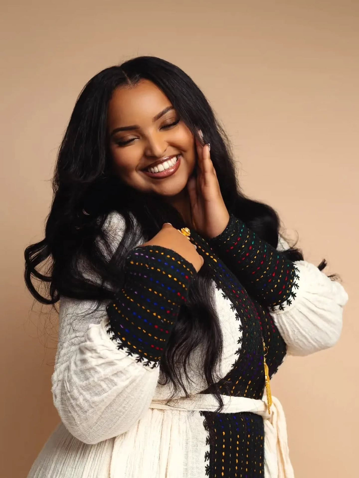 A smiling woman with long black Habesha Kemis poses in a white textured top and a black embroidered cardigan with multicolored dotted accents, set against a solid beige studio background.
