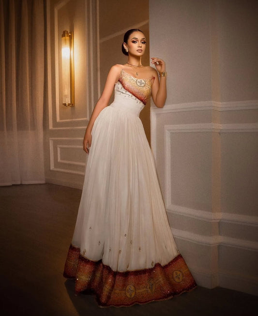 A woman wearing a strapless white tulle Habesha Dress  with an ornate gold and red embroidered bodice, posing against a gray paneled wall.