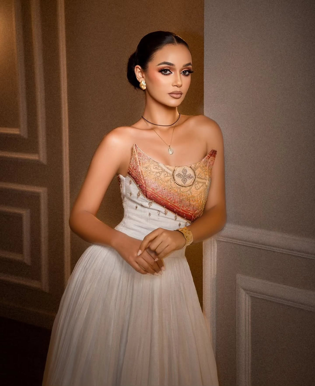 A woman wearing a strapless white tulle Habesha Dress  with an ornate gold and red embroidered bodice, posing against a gray paneled wall.