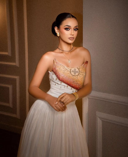 A woman wearing a strapless white tulle Habesha Dress  with an ornate gold and red embroidered bodice, posing against a gray paneled wall.