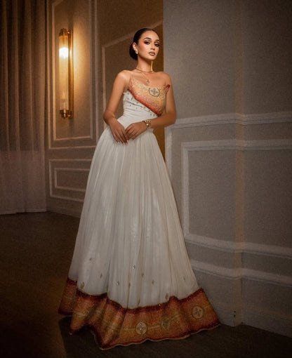 A woman models a strapless white Habesha Dress  with gold and orange-red embroidered details on the bodice and a wide decorative border at the hem, set against an elegant indoor hallway with warm wall lighting.