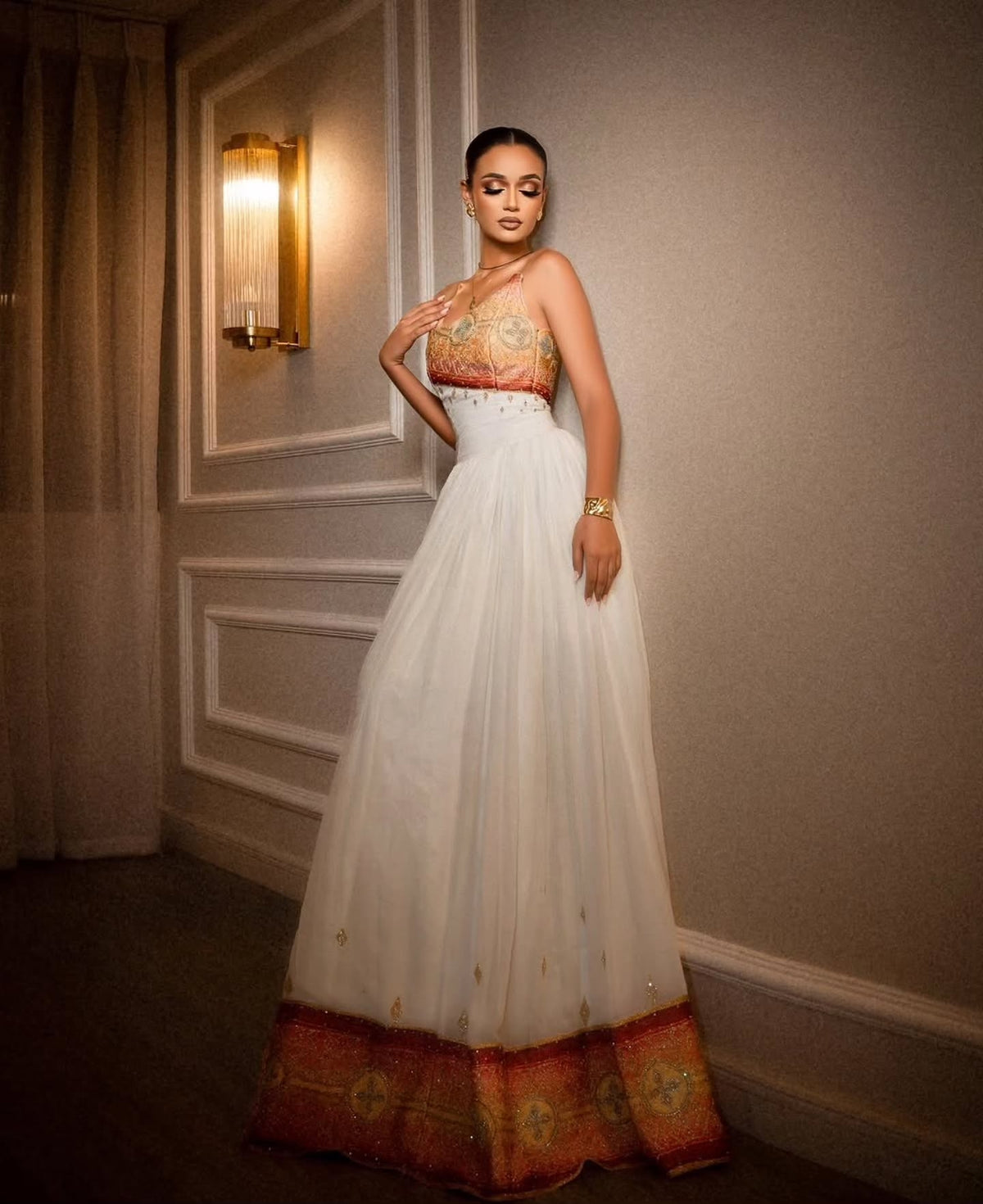 A woman wearing a strapless white tulle Habesha Dress  with an ornate gold and red embroidered bodice, posing against a gray paneled wall.
