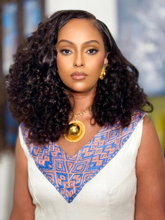 A woman with voluminous dark curly hair is shown from the chest up, wearing a white habesha kemis with a blue and orange geometric patterned V-neckline, a large gold pendant necklace, and gold earrings.