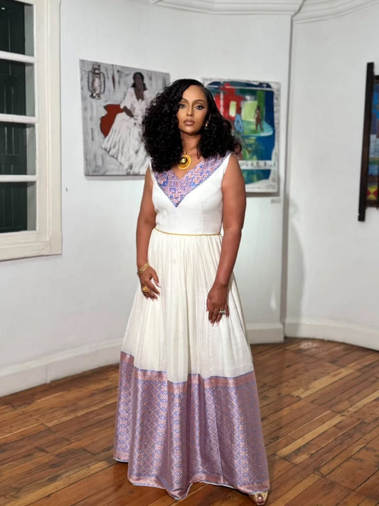 A woman stands in a room with white walls and wood floors, wearing a sleeveless white habesha kemis with purple patterned trim at the neckline and a matching purple patterned hem, along with gold jewelry.