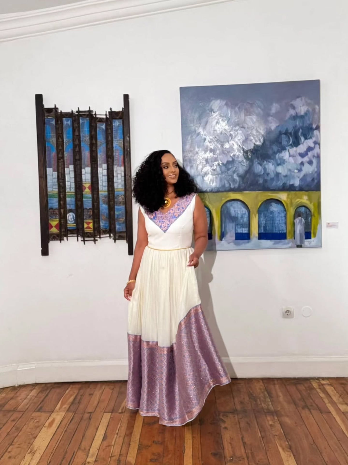 A woman wearing a sleeveless white and lavender habesha kemis stands in a gallery room with wood floors, posing in front of a colorful painting and a stained glass window.