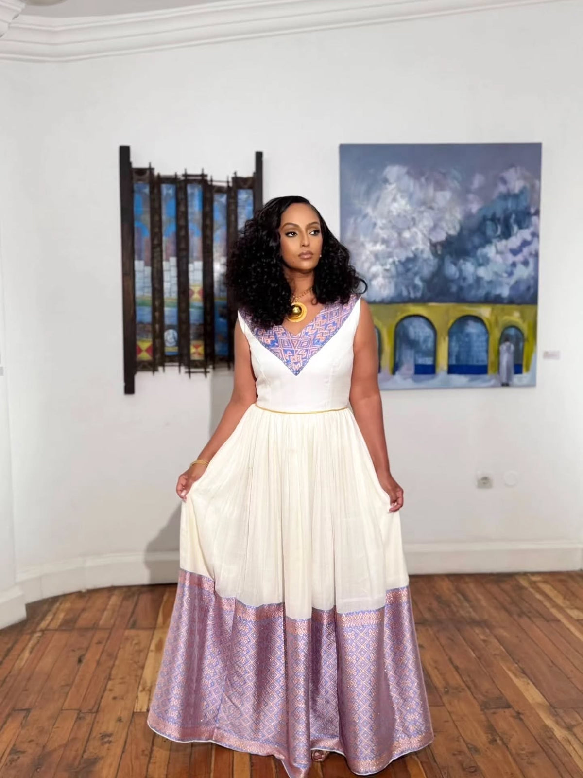 A woman modeling a sleeveless habesha kemis with a white upper bodice and a lavender patterned skirt, standing in a gallery with paintings on the wall and a wooden floor.