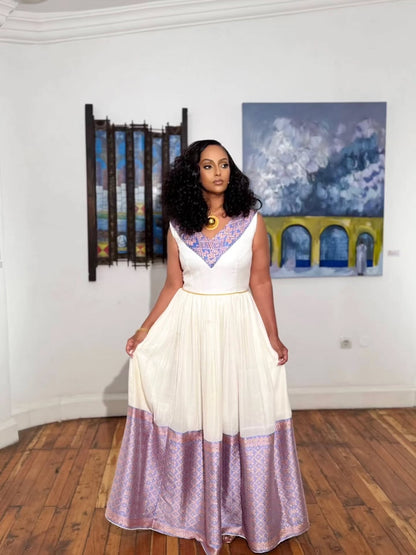 A woman modeling a sleeveless habesha kemis with a white upper bodice and a lavender patterned skirt, standing in a gallery with paintings on the wall and a wooden floor.