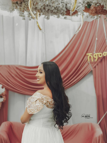 Woman in a white Gold Habesha Kemis with lace details standing in front of a decorated backdrop with red curtains and floral arrangements.