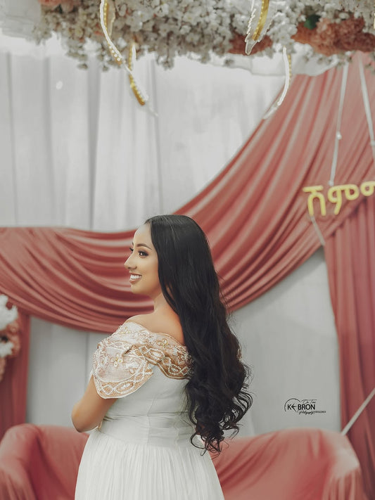 Woman in a white Gold Habesha Kemis with lace details standing in front of a decorated backdrop with red curtains and floral arrangements.