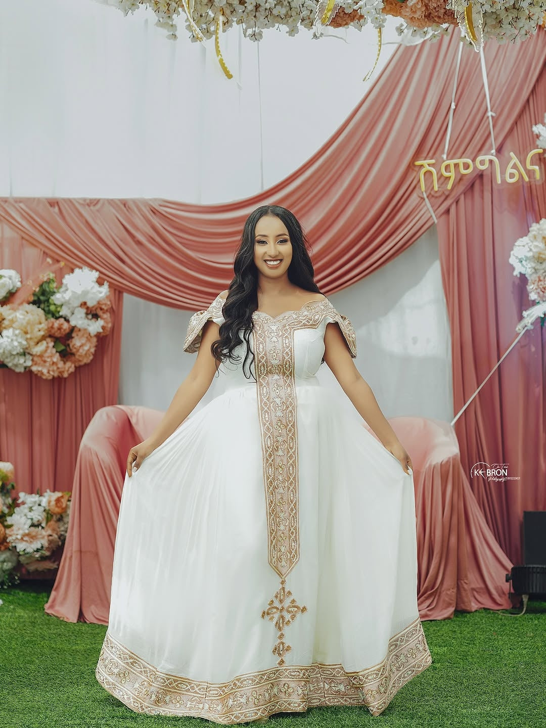 Woman in a white and gold Gold Habesha Kemis standing in front of a decorated backdrop with pink drapes and floral arrangements.