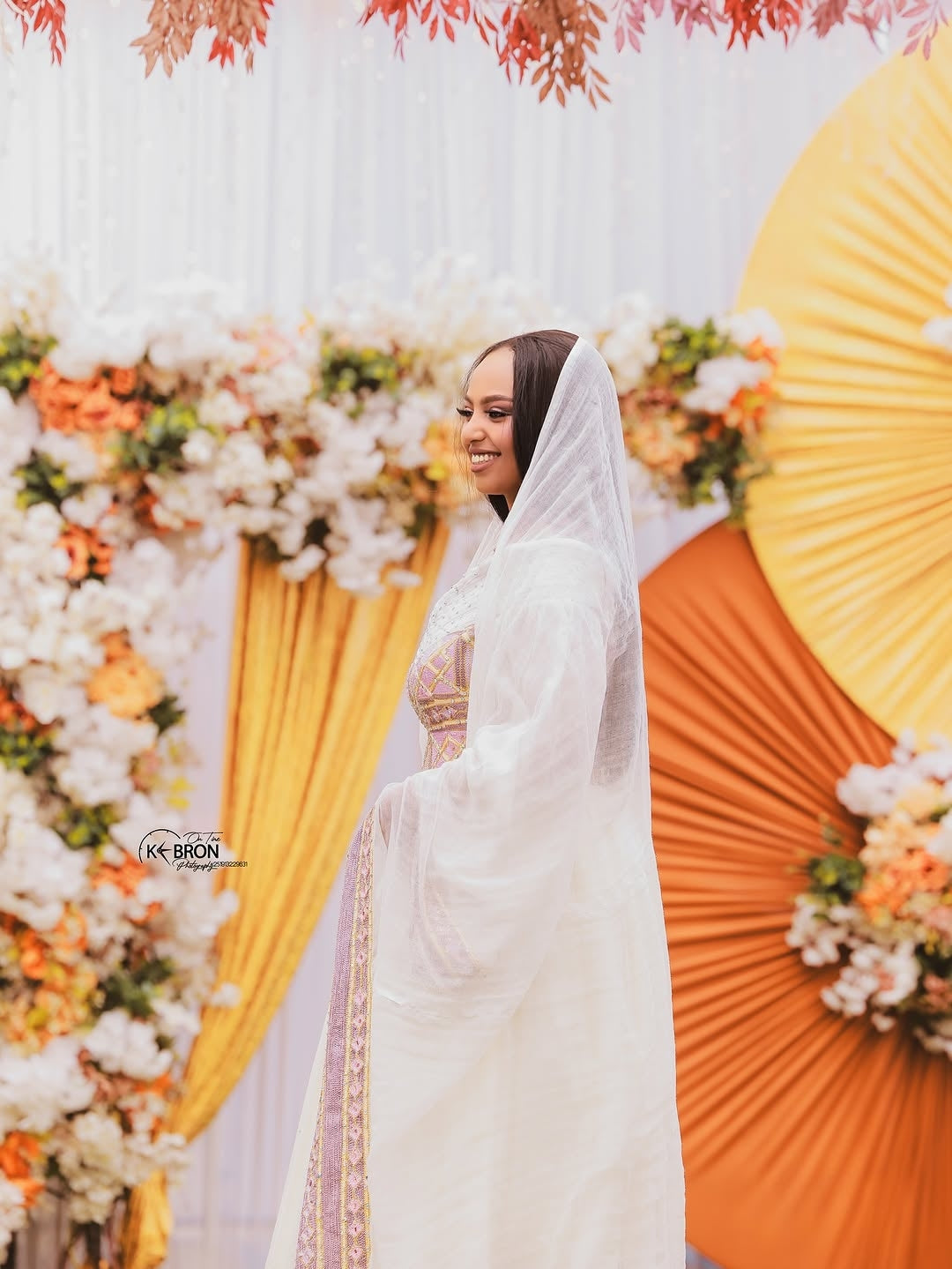 Woman in a white veil standing in front of a Ethiopian Dress  floral backdrop with yellow and orange elements.