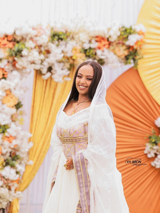 Woman in a white and gold traditional outfit standing in front of a floral and draped backdrop.