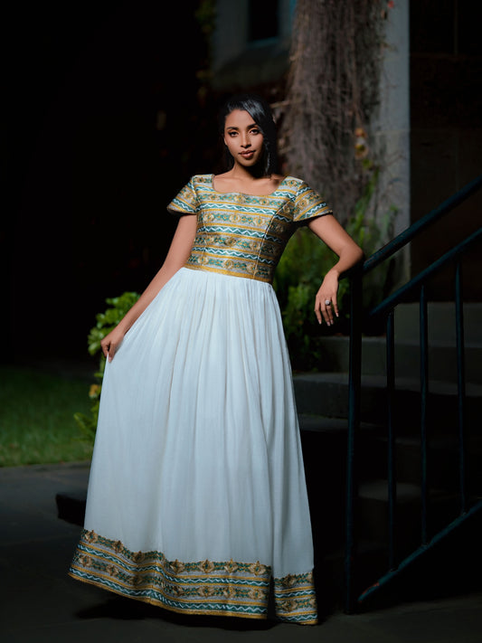 A woman wearing a floor-length white Green Habesha Kemis with a gold and teal patterned bodice and hemline, standing on outdoor stairs at night.