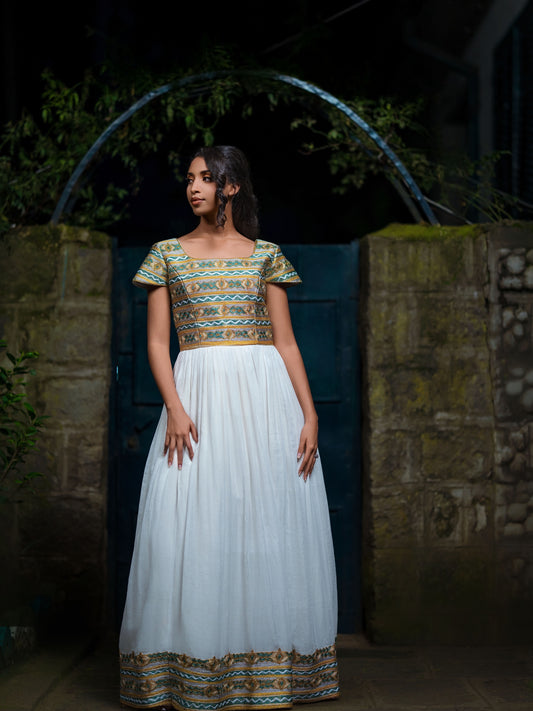 A woman stands in front of a dark blue door framed by stone walls at night, wearing a long white Green Habesha Kemis with a heavily embroidered gold, green, and blue bodice and border.