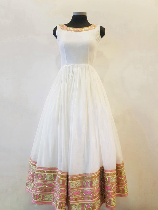 A sleeveless white Anarkali-style dress displayed on a black Ethiopian Outfit against a plain light beige wall. The dress features a fitted bodice, a full flared skirt, and ornate pink and gold embroidered borders at the neckline and hem.