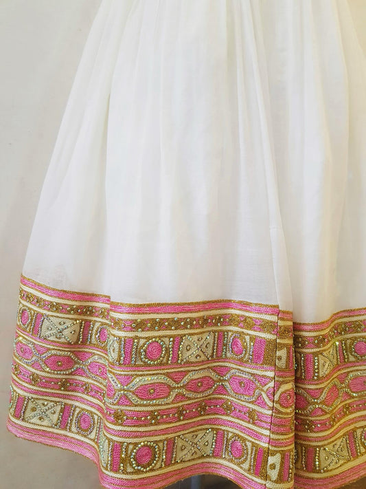 Close-up of a white Ethiopian outfit with a wide ornate border featuring pink, gold, and beige embroidered patterns.