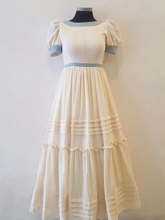 A cream-colored tiered Ethiopian Outfit with puff sleeves and light blue trim on the neckline, sleeve cuffs, and waist, displayed on a black mannequin against a plain off-white wall.